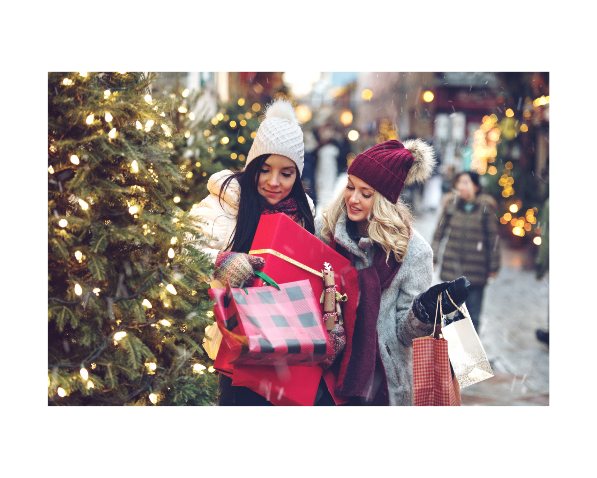 Zwei Frauen sind beim Einkaufen in der Weihnachtszeit unterwegs. Sie tragen Mützen und jeweils viele Einkaufstaschen aus dem lokalen Handel. Ein Weihnachtsbaum schafft Atmosphäre.